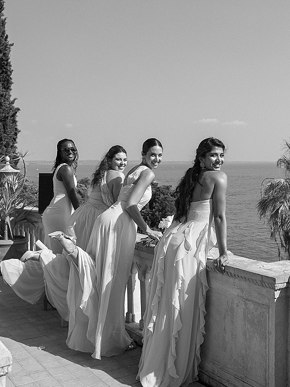 Bridesmaids portrait of a group laughing and looking back on a stone terrace with ocean and sky behind, holding bouquets and wearing sunglasses