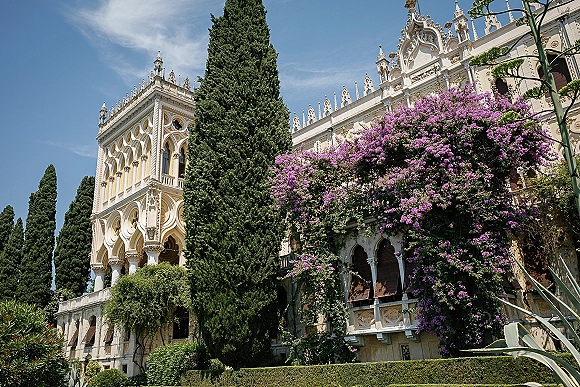 Wedding venue exterior with climbing bougainvillea on an ornate historic facade, framed by cypress trees, hedges, and blue sky