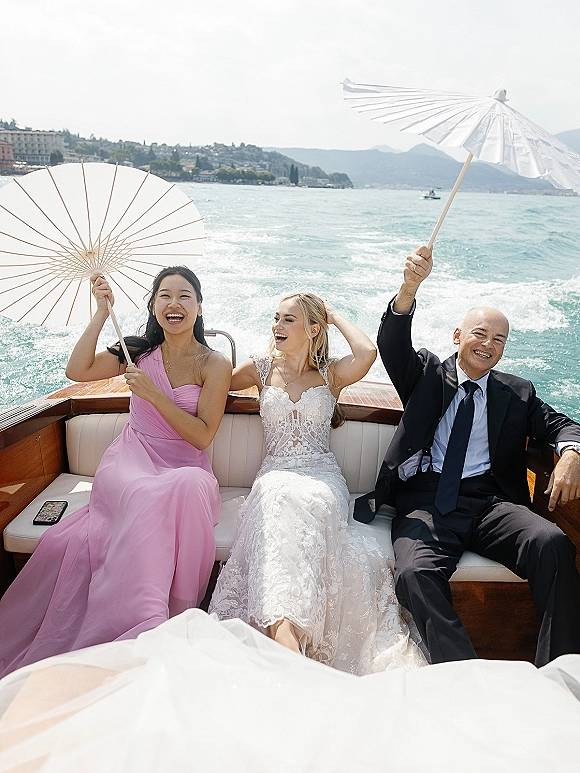 Wedding boat portrait of bride in lace dress and veil with groom in black suit and wedding party on a speedboat, mountains behind