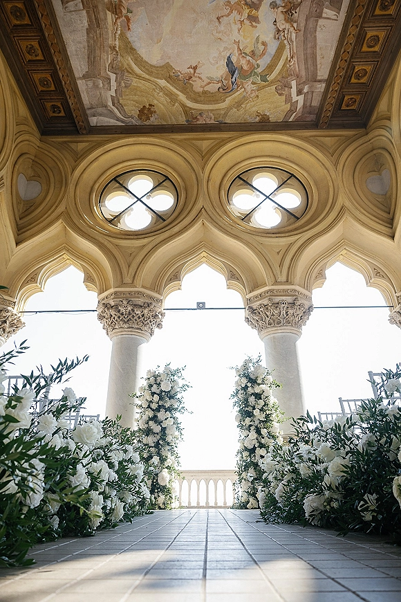 Ceremony aisle decor with white floral arrangements and greenery garlands lining a runner beneath an arched colonnade and fresco ceiling mural