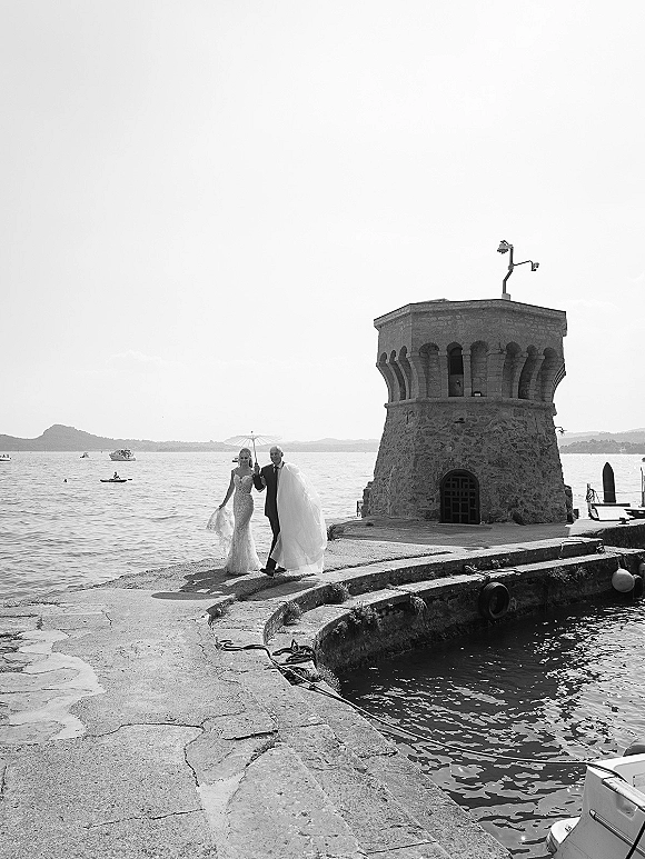 Couple portrait in a black and white wedding photo, bride and groom walking on a pier with parasol, stone tower and boats behind them
