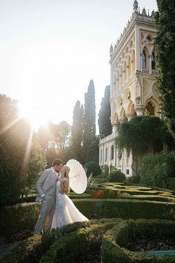 Wedding couple portrait of bride and groom kissing under a paper parasol, backlit by sun flare in a formal garden by a historic building