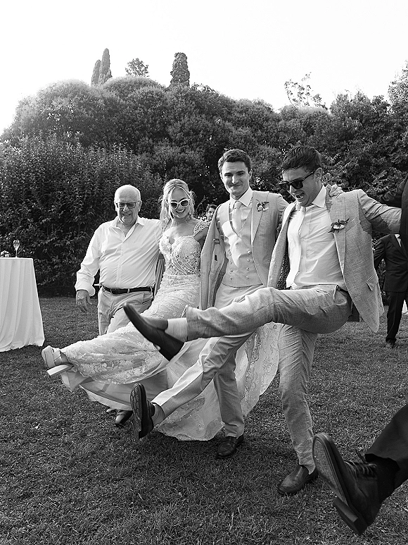Wedding group portrait of the bridal party walking and kicking legs, bride in lace dress with sunglasses on a garden lawn with hedges