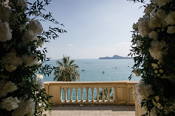 Ceremony arch draped in a wedding floral arch of white flowers and greenery, set on a terrace with ocean view and boats beyond