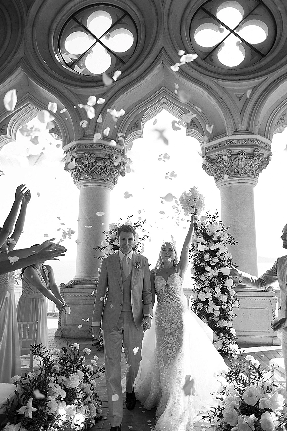 Wedding recessional as bride and groom hold hands walking down the aisle, bride lifting bouquet overhead amid flower petal toss on terrace