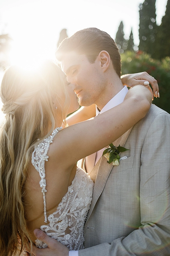 Wedding kiss portrait of bride and groom kiss, her lace dress and ring visible as sun flare glows through garden trees