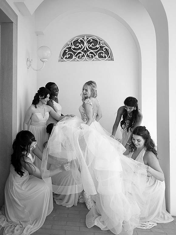 Bride getting ready as bridesmaids help bustle her lace bodice tulle skirt in an arched white hallway with tile floor and wall sconce