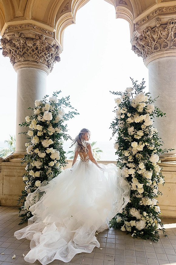 Bridal portrait of a bride in a backless wedding dress with cathedral train, posing over her shoulder by floral pillars at a stone arch terrace