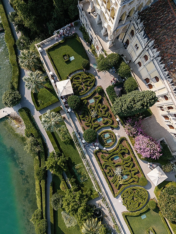 Wedding venue aerial view of a formal garden parterre with manicured hedges, gravel paths, and white patio umbrellas by a lakeside estate