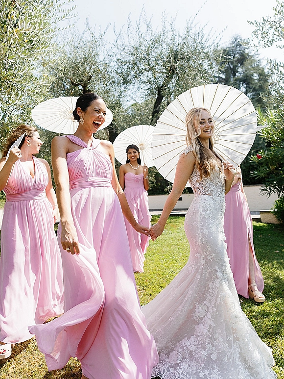 Bride with bridesmaids walking hand in hand in pink bridesmaid dresses, holding parasols on a sunny garden lawn with trees and blue sky