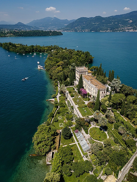 Wedding venue aerial showing a historic villa with formal gardens and topiary hedges on a lakefront terrace with dock and boats, mountains beyond