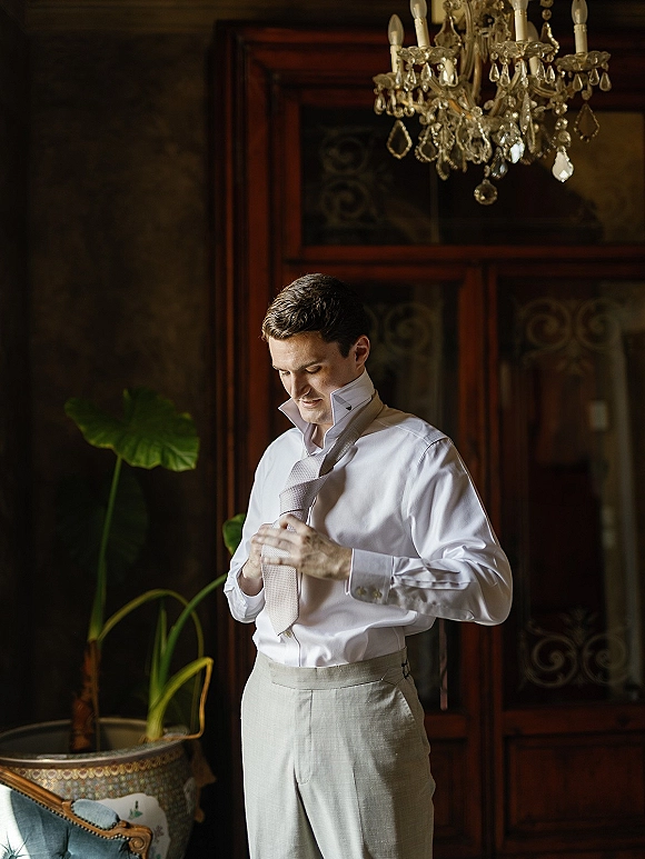 Groom getting ready, groom tying tie in a dark vintage room with patterned wall, wearing a white dress shirt and cufflinks under a chandelier
