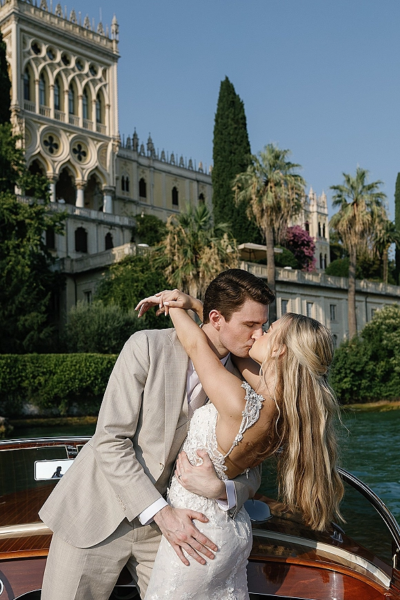 Wedding kiss portrait of newlyweds kissing on a speedboat, bride in beaded lace straps and rings visible against palm-lined waterfront villa backdrop
