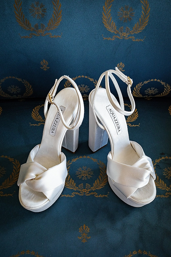 Bridal shoes, white wedding heels with ankle straps and knotted satin toe straps, resting on blue upholstery with gold floral motifs