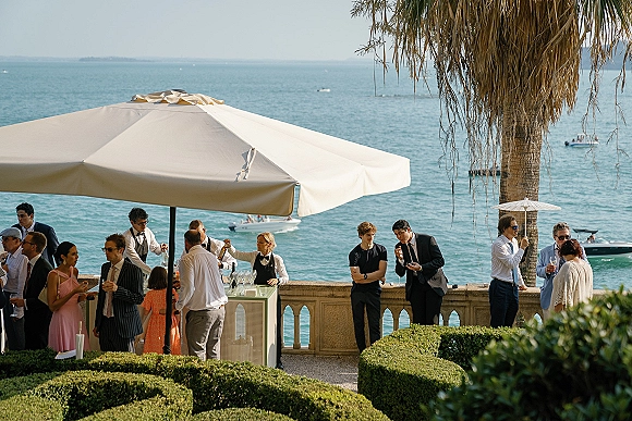 Wedding cocktail hour at an outdoor wedding bar with bartenders pouring champagne as guests mingle on a seaside stone terrace under umbrellas