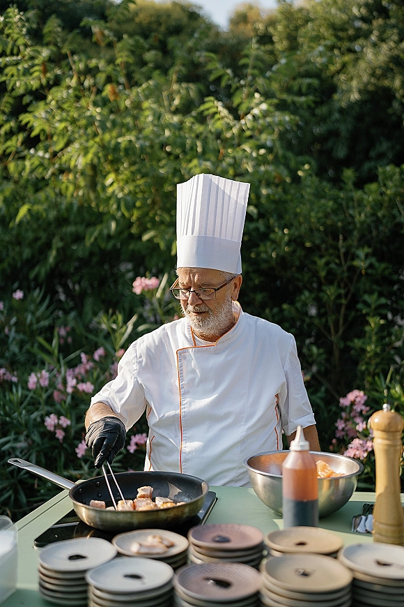 Wedding catering chef at a live wedding cooking station, wearing tall hat and black gloves, frying appetizers outdoors amid garden flowers