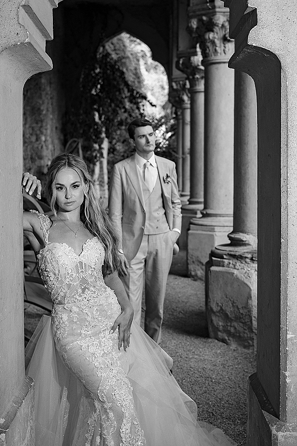 Couple portrait in black and white with bride in lace corset dress leaning on a stone archway as groom stands behind her by columns