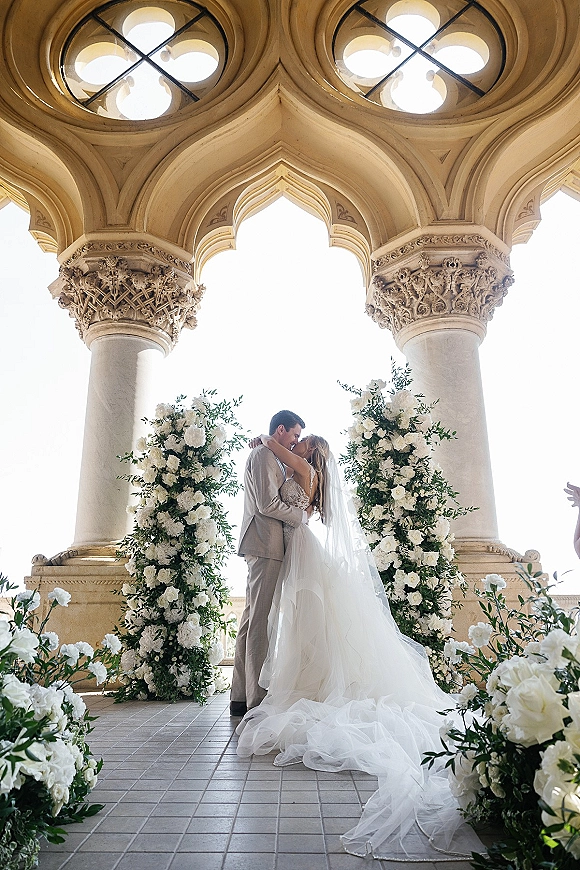 Wedding kiss portrait of bride and groom kissing with a long veil accent between white floral pillars on an ornate stone-arched terrace