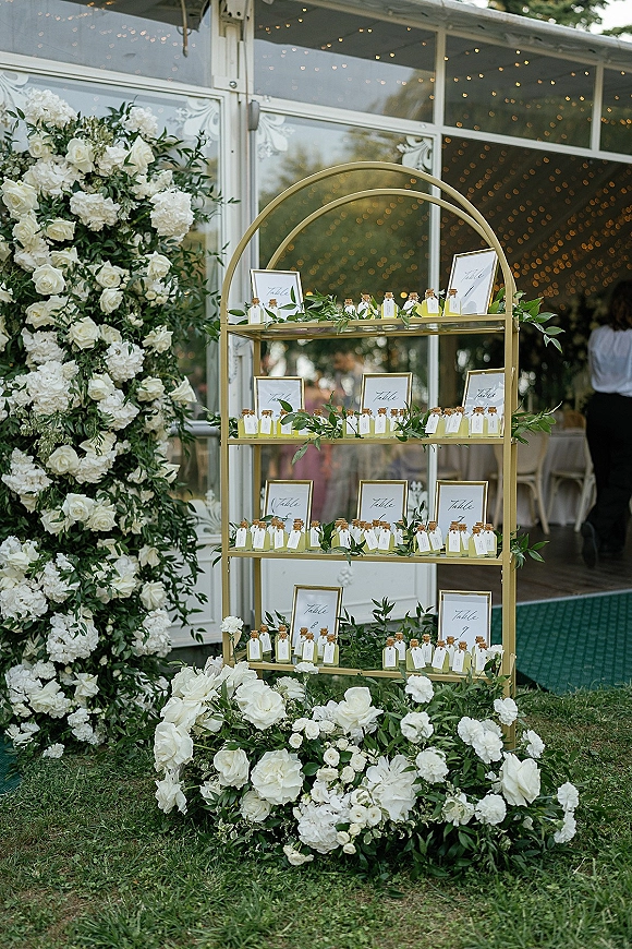 Wedding seating chart on gold shelving display with framed escort cards and white rose and hydrangea greenery under string lights by a glass wall