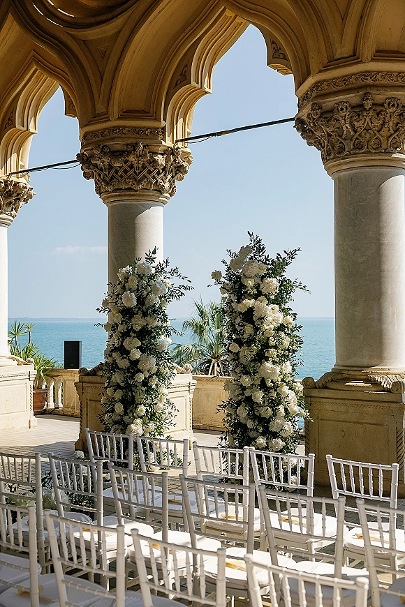 Ceremony setup with white floral pillars and greenery lining an aisle runner, white chiavari chairs on a stone terrace overlooking the ocean.