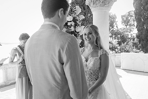 Wedding vows as bride in lace dress and veil smiles at groom in gray suit under floral arch on ocean-view terrace with columns