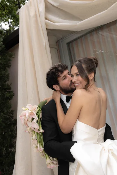 Couple portrait of the groom kissing the bride on the cheek as she holds a calla lily bouquet, framed by draped fabric and a sheer-curtained window.