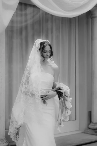 Bridal portrait in black and white, bride with a lace veil gazing down, framed by columns and sheer curtains near a doorway.