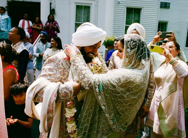 Ceremony moment at a Sikh wedding ceremony as the bride places a white floral garland on the groom in a turban, guests filming outside a house