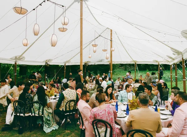 Wedding reception tent with string lights and paper lanterns over round tables set with floral centerpieces, blue bottles, and bentwood chairs on a lawn