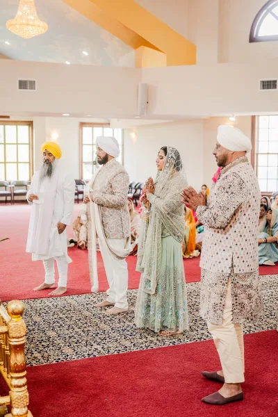 Wedding ceremony moment as Sikh bride and groom stand with hands in prayer on a red carpet beneath a chandelier in an indoor hall