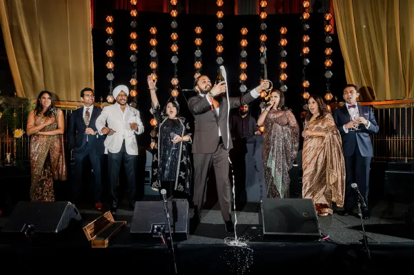 Wedding champagne pop as guests in suits and sarees cheer on a stage, champagne spray bursting under stage lights against gold curtains