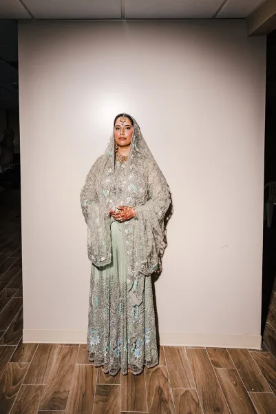 Bridal portrait of a mint green lehenga bride with dupatta veil, maang tikka, and mehndi hands, standing by a plain indoor wall