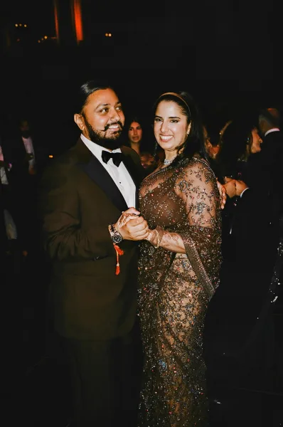 Couple portrait at wedding reception, bride in sequined gown holding hands with groom in black tuxedo, smiling in warm dim room with guests