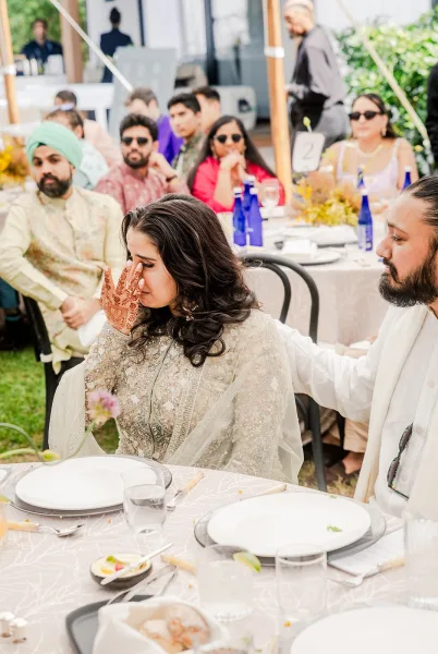 Wedding reception moment as the bride wipes tears with mehndi hands, embroidered dupatta draped at an outdoor tented table setting with floral centerpiece