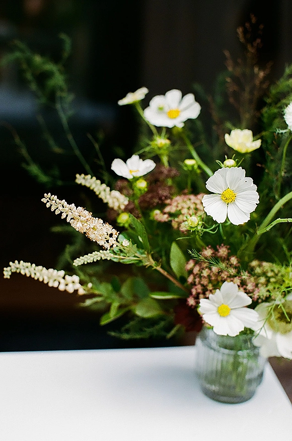 Wedding centerpiece with wildflower wedding centerpiece blooms, white cosmos and greenery in a glass jar vase on a white tablecloth, dark blurred backdrop