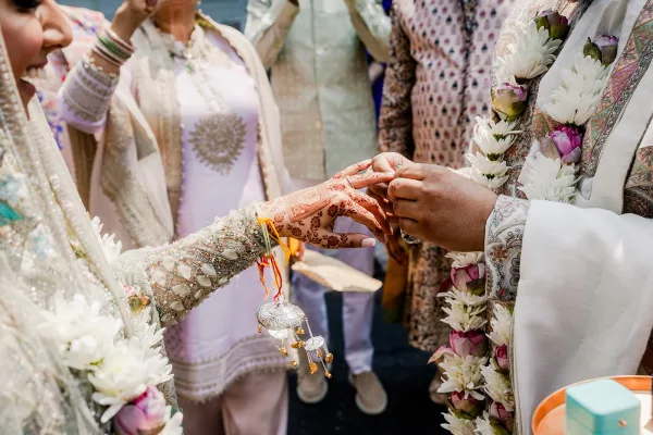 Ring exchange as groom places wedding ring on bride’s hennaed finger, bangles and floral garlands visible, guests watching outdoors