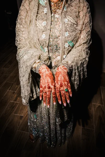 Bridal mehndi hands with henna hand designs, ring and neutral nails, framed by embroidered dupatta and necklace in a dark hallway