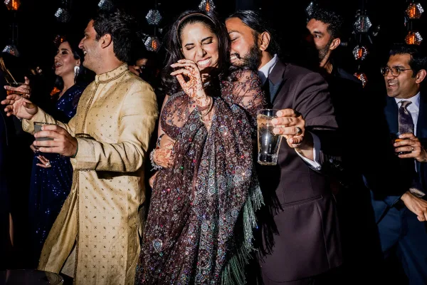 Wedding dance floor packed with guests dancing under hanging lantern lights, sequined outfits and sherwani, with hennaed hands holding cocktails