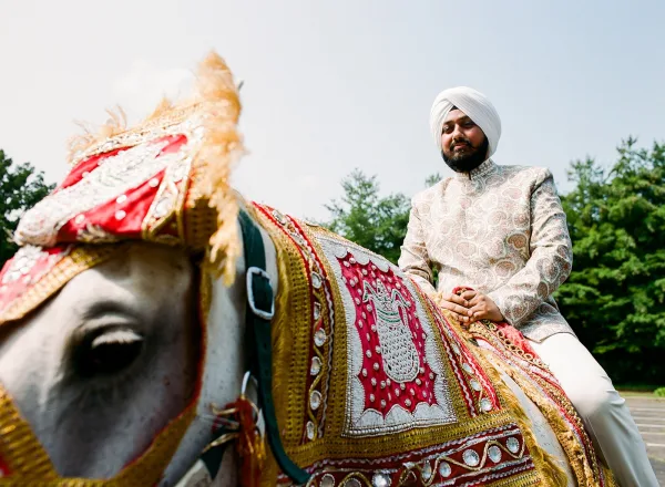 Groom baraat entrance with groom on horse in an embroidered sherwani and white turban, riding under blue sky along a tree-lined road