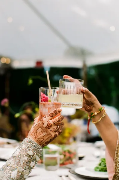 Wedding toast with henna hands raising cocktail glasses and straws, bangles and embroidered sleeve at a tent reception under bistro lights