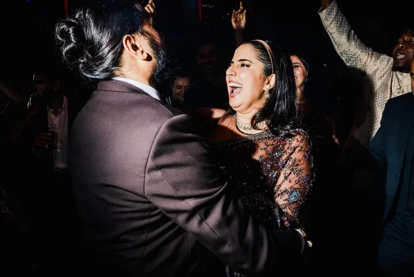 Wedding dance moment as couple dances in a cheering reception crowd under dim lighting, bride in sparkly black evening gown with statement earrings