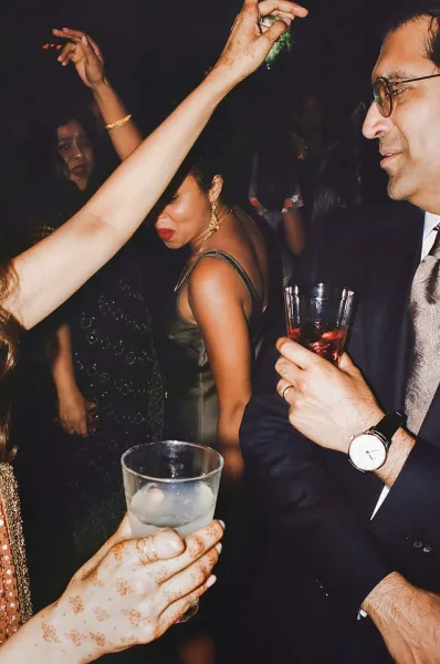 Wedding dance floor reception dancing with guests raising cocktail glasses, henna hands with gold bangles visible in a dark party room