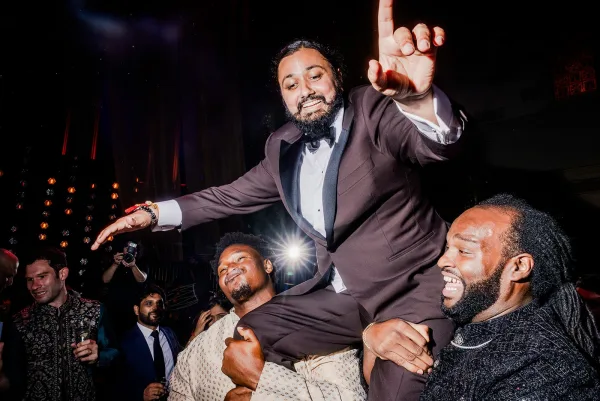 Wedding dance floor scene with groom in tuxedo lifted in a chair by friends, bow tie and wristwatch visible as guests cheer in dark reception lighting