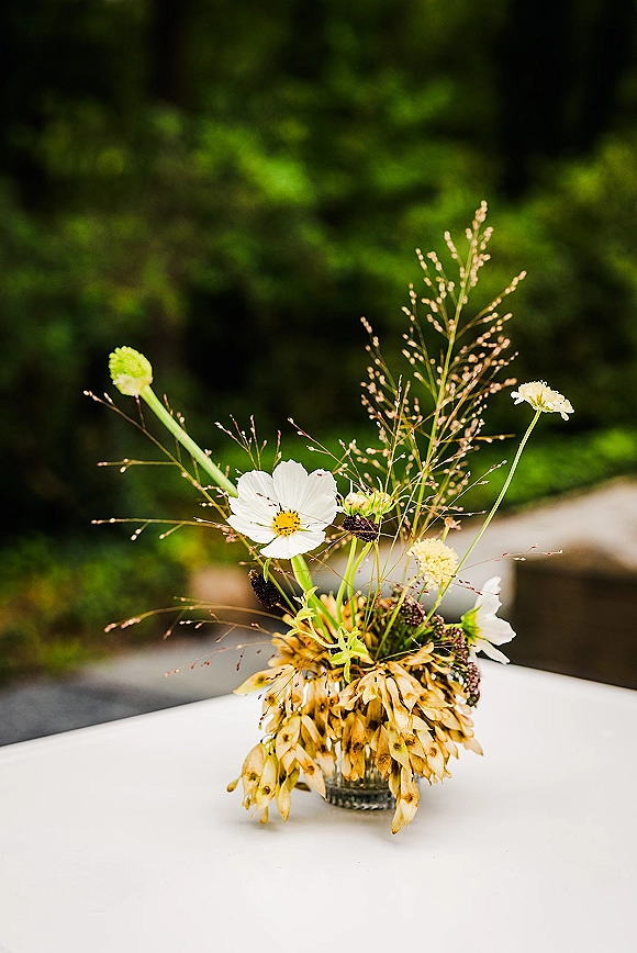 Wedding centerpiece of white wildflowers and dried pods in a glass jar vase on a white tablecloth with blurred outdoor greenery behind