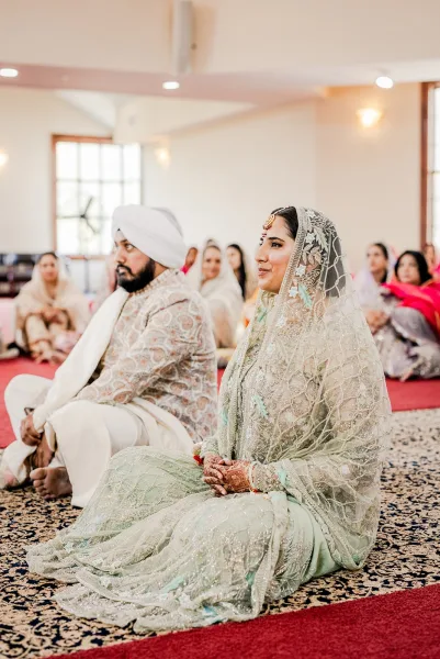 Ceremony moment at a Sikh wedding ceremony with bride and groom seated in lehenga and turban on a patterned carpet in an indoor hall