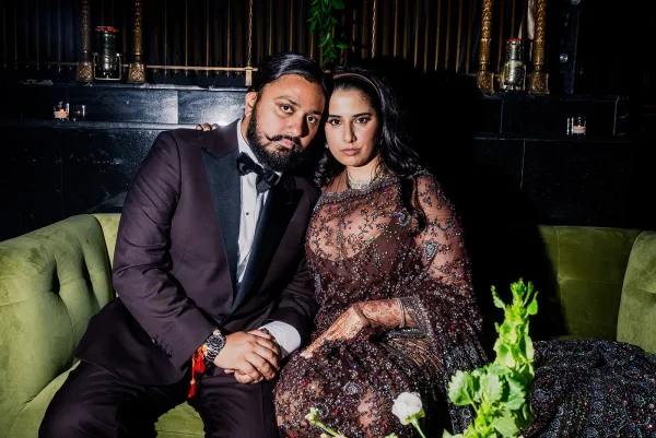 Couple portrait of bride in a beaded gown with henna and statement jewelry beside groom in tuxedo on a velvet sofa in a dark lounge with candles