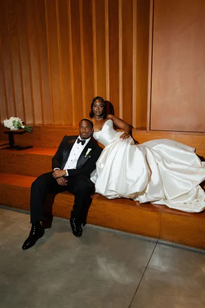 Couple portrait with a bride in a strapless gown beside her groom, seated on wooden steps against a wood panel wall in an editorial pose.