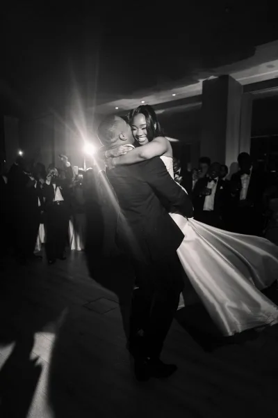 Wedding first dance with the groom lifting the bride under a warm spotlight on the indoor reception dance floor, his classic tuxedo crisp.