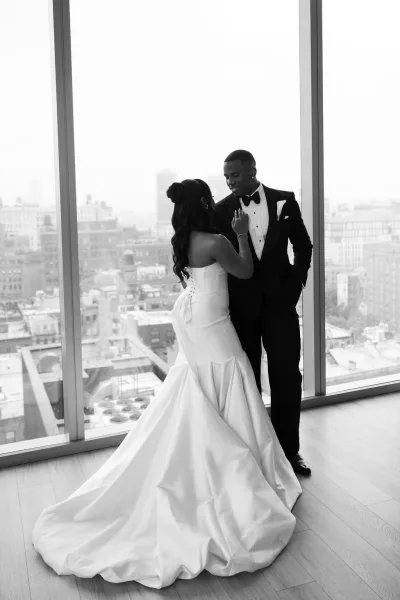 Couple portrait in black and white, bride adjusts the groom’s bow tie, framed by large windows with a sweeping city skyline.