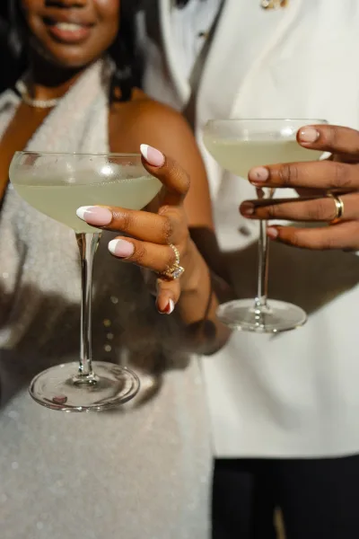 Champagne toast as a couple holding cocktails clink coupe glasses, framed by sparkly fabric and a white blazer, capturing an intimate wedding moment.
